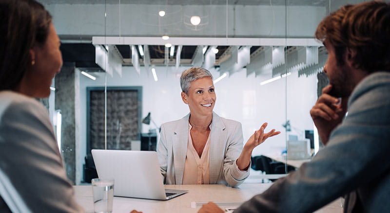 Person with laptop talking with hands to two others in office