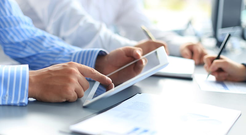 Three sets of arms and hands working on a tablet, notepad and papers with charts and graphs