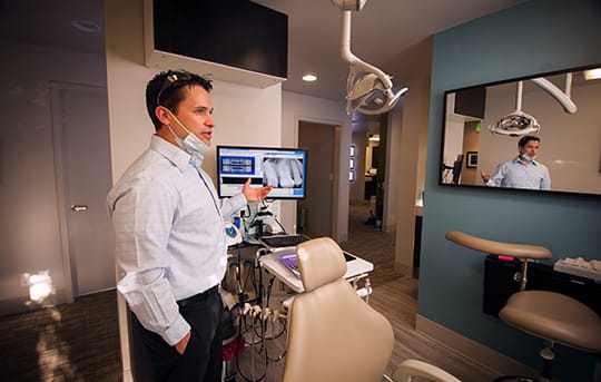 Dentists office with dentist gesturing toward display screen with tooth x-ray image
