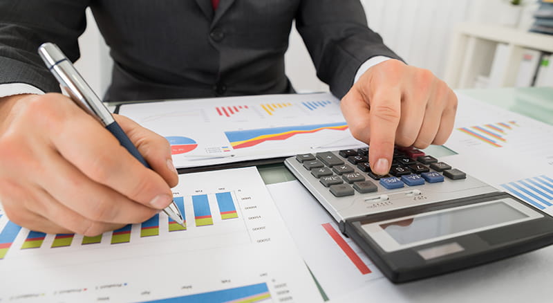Man's hands holding pen and using a calculator with papers of charts and graphs