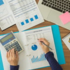 Feminine hands holding a pencil and keying a calculator with papers underneath that has graphs