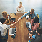 Six people conducting a business meeting at a long wooden table, two people shaking hands across the table