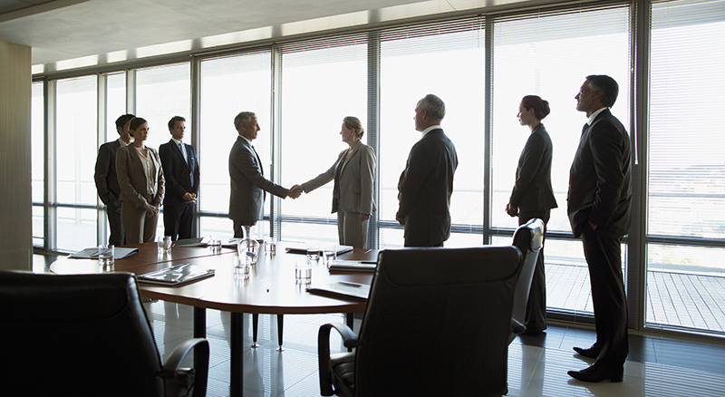 Group meeting in a large conference room two people in the center shaking hands as if two groups are making a deal