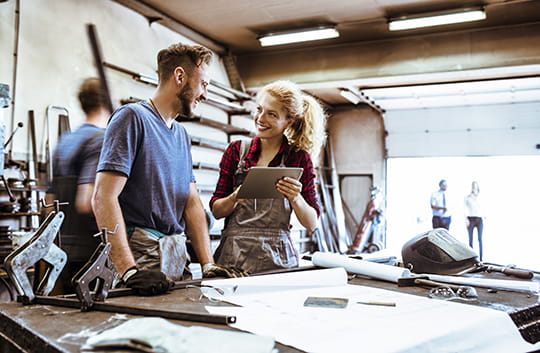 White man with brown hair and beard speaking with a blonde haired white woman standing at a work table inside a big manufacturing shop