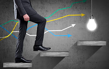 Lower half of man in business attire walking up stairs with lines graph in the background walking up stairs toward a light bulb