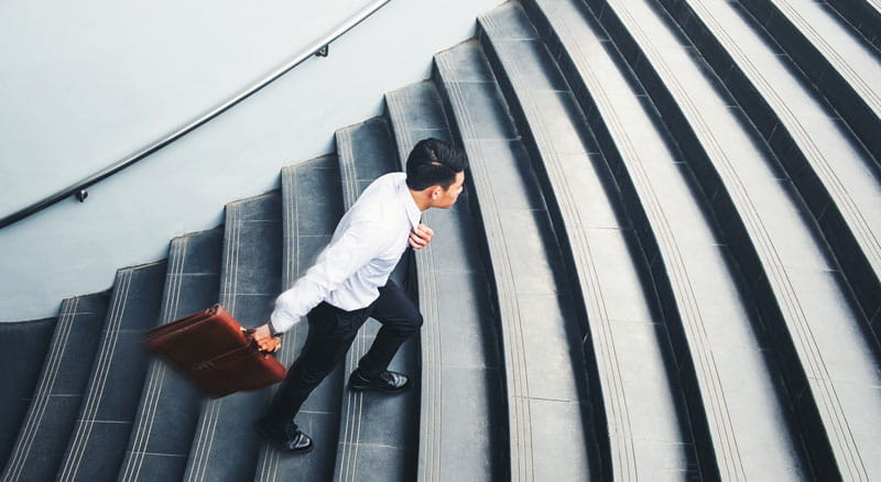 Asian man, mid-step, in business attire walking up cement steps carrying a brown leather briefcase