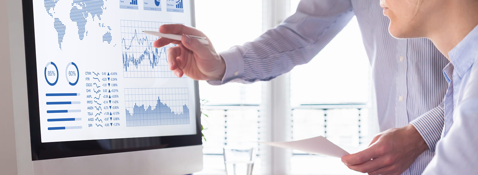 Two business people looking at a large computer screen with charts and graphs one person pointing at screen with a pen and second person seated looking at screen