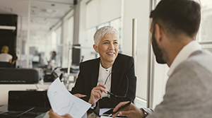 Business woman with short white hair speaking with a dark haired and bearded business man with his side and back to the camera