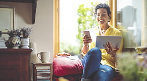 Women sitting by a window holding a tablet and looking at her smartphone