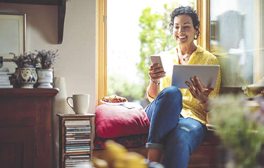 Women sitting by a window holding a tablet and looking at her smartphone.