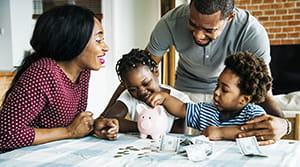 African American mom and dad with son and daughter at a table with cash and coins surrounding a pink piggy bank