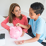 Mother teaching son about savings, boy inserting coins in pink piggy bank