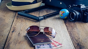 Travel essentials laid out on a wooden table - hat, sunglasses, camera, sunglasses, passport and foreign currency