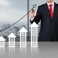 Business man in suit draws upward arrow above model buildings that increase in height