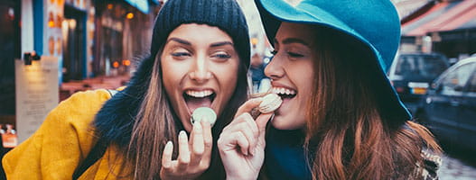 Two women in hats and coats laugh while eating macarons