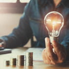 Ascending stacks of coins with a lit clear light bulb at the end next to the tallest stack the light bulb is being held by a man in a denim blue shirt