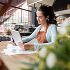 Dark haired female shop owner holding a tablet and stacks of papers next to her