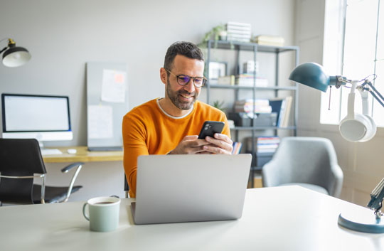 Office worker looks at mobile phone while working on laptop